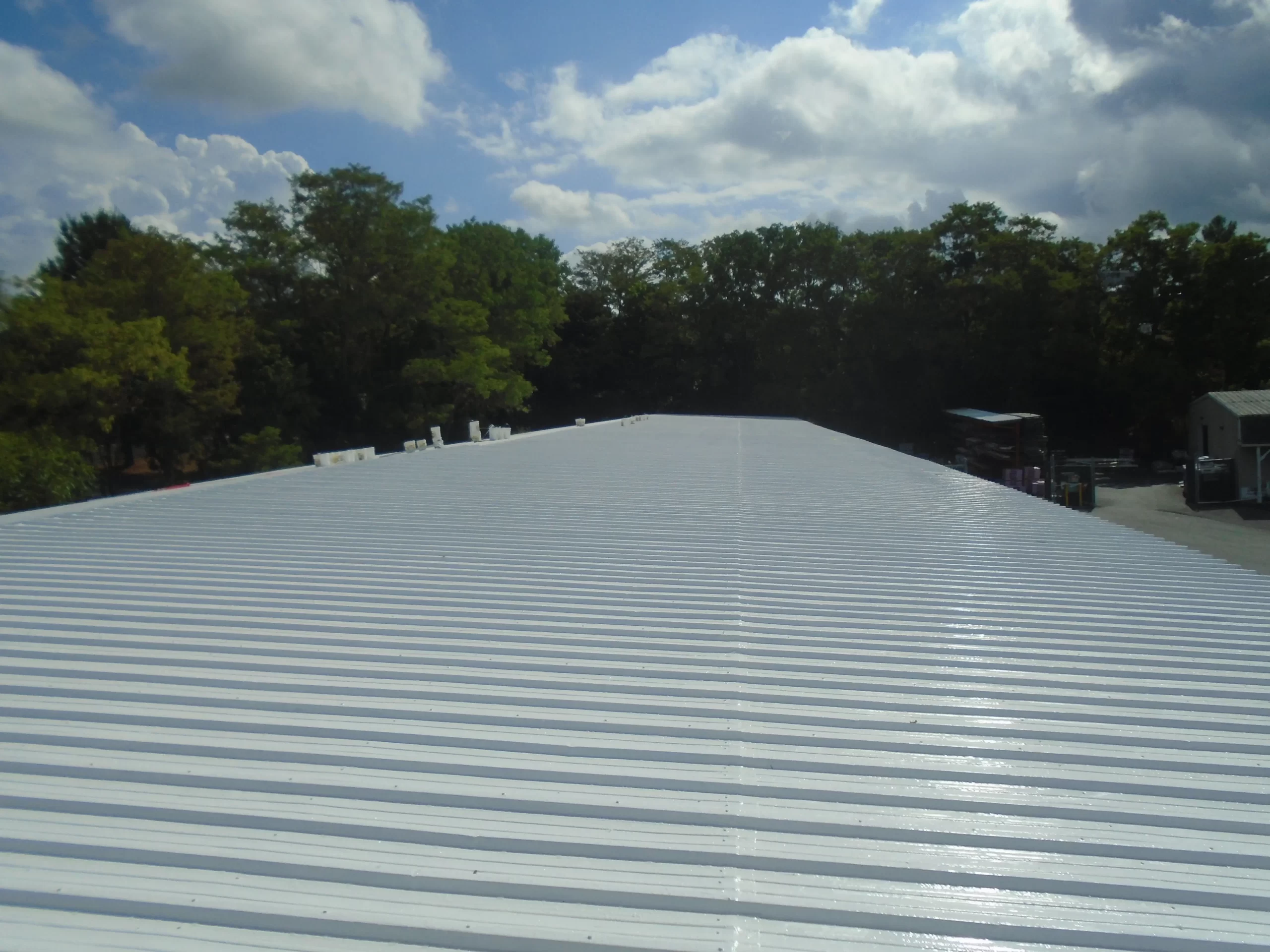 A freshly coated white metal roof in Danbury, CT, reflecting sunlight with trees and an industrial yard in the background.