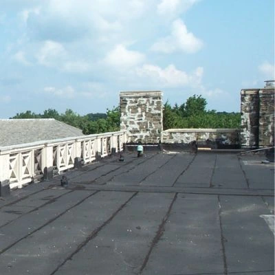 Commercial flat roof with modified bitumen membrane roofing system, stone chimneys, and decorative balustrade against a blue sky background.