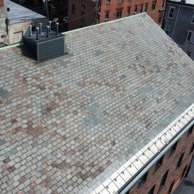 Aerial view of an aging tile roof on an urban building with visible weathering patterns and HVAC equipment, surrounded by brick apartment buildings.